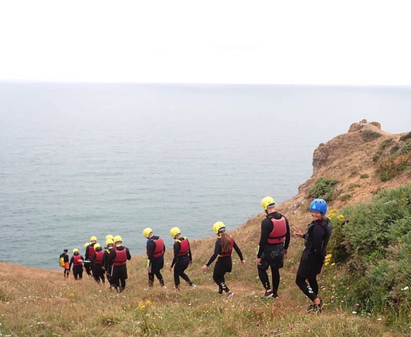 Family coasteering group in wetsuits and helmets walking down the clifftop path to the sea near Tenby